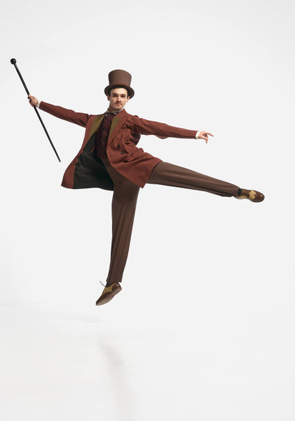 Levitating body. Portrait of an attractive man wearing old clothes and black cylinder jumping with cane up over white studio background. Concept of historical remake, comparison of eras, vintage - Foto, immagini