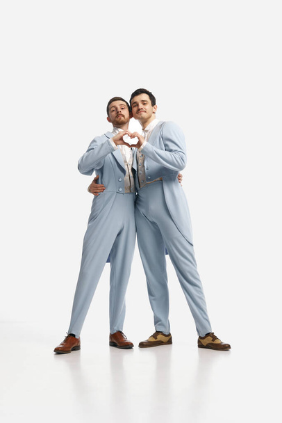 Shot of two handsome young men wearing blue suit standing together and showing heart with hands. Human relationship. Concept of lgbt, love, gay, rights, comparison of eras, retro, vintage, emotions - Foto, Imagen