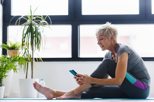 mujer mayor sentada en esterilla de yoga con teléfono  - Foto, imagen
