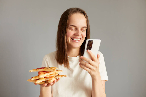 Portrait of joyful woman with pizza in hands, enjoying fast food isolated over gray background using mobile phone leaves positive feedback to the delivery service and pizzeria. - Photo, Image