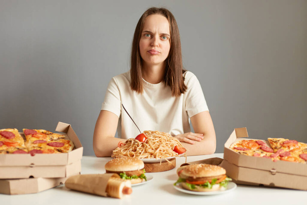 Portrait of upset irritated woman with brown hair wearing white T-shirt sitting at table isolated over gray background, being dissapointed of junk food menu, prefers healthy eating. - Foto, Bild