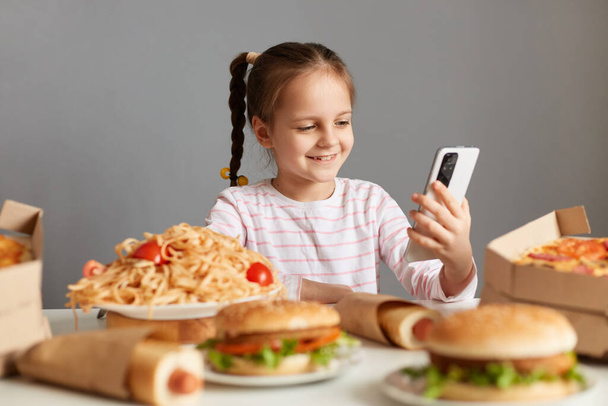 Photo of cute cheerful hungry little girl with pigtails sitting at table isolated over gray background, having video call, saying about her cheat meal, being surrounded with different junk food. - Foto, Imagen