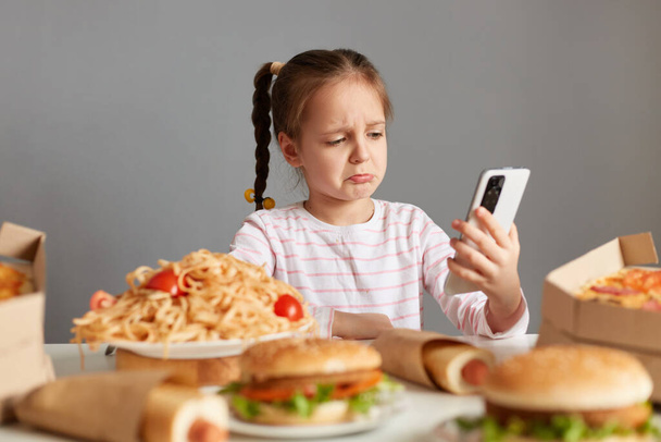 Indoor shot of sad crying little girl with pigtails sitting at table pizza, hot dogs, burgers, talking via video call, keeps diet, can't eat delicious fast food, isolated over gray background - Photo, Image