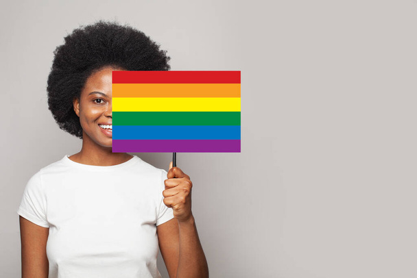 Portrait d'une femme afro-américaine souriante et heureuse tenant une pancarte avec un drapeau LGBT sur fond gris - Photo, image