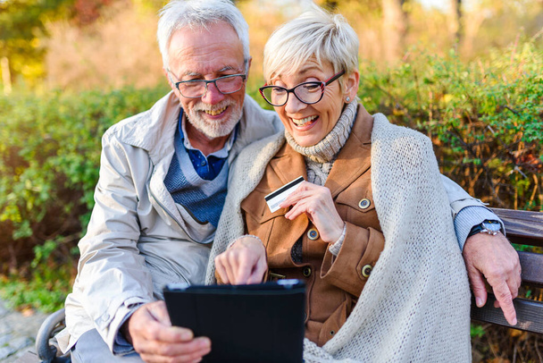 ηλικιωμένο ζευγάρι κάθεται στον πάγκο στο πάρκο, κάνοντας online παραγγελία - Φωτογραφία, εικόνα