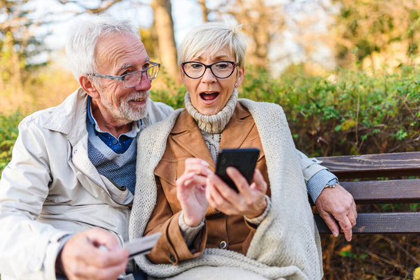 ηλικιωμένο ζευγάρι κάθεται στον πάγκο στο πάρκο, κάνοντας online παραγγελία - Φωτογραφία, εικόνα
