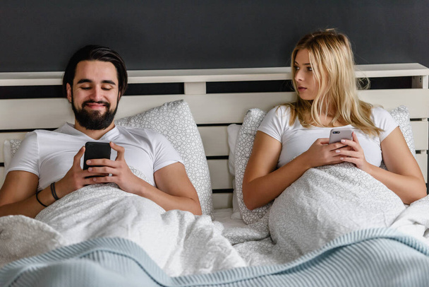 üzgün genç bir çift yatakta uzanıyor ve akıllı telefonlar kullanıyor. - Fotoğraf, Görsel