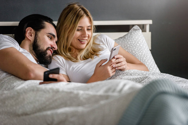 νεαρό ζευγάρι που χρησιμοποιεί τηλέφωνα στο κρεβάτι στο σπίτι - Φωτογραφία, εικόνα
