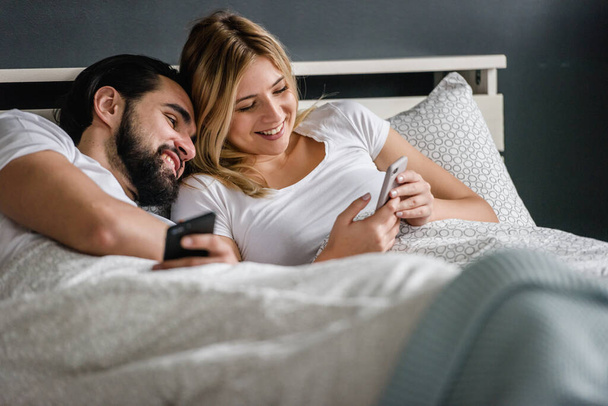 happy couple lying in bed and looking at smartphone in morning - Photo, Image