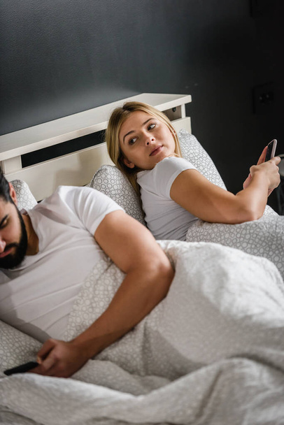 üzgün genç bir çift yatakta uzanıyor ve akıllı telefonlar kullanıyor. - Fotoğraf, Görsel