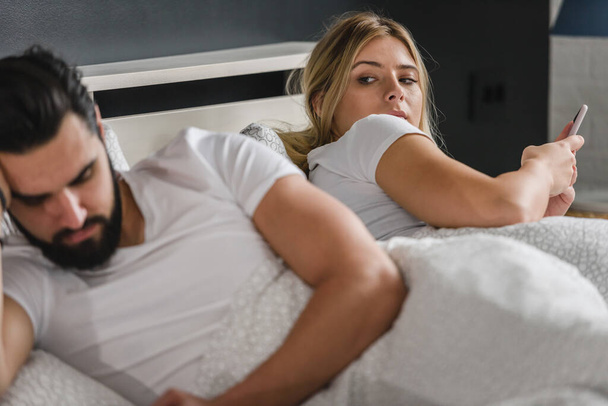 αναστατωμένο νεαρό ζευγάρι ξαπλωμένο στο κρεβάτι και χρησιμοποιώντας smartphones - Φωτογραφία, εικόνα
