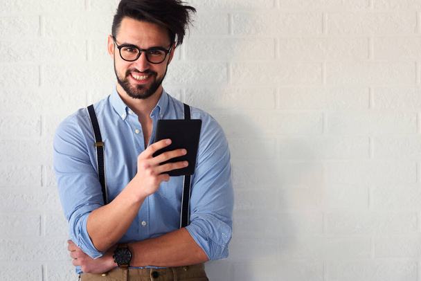 pohledný mladý muž s vousy a brýlemi drží tablet a usmívá se, zatímco stojí u zdi a dívá se jinam - Fotografie, Obrázek