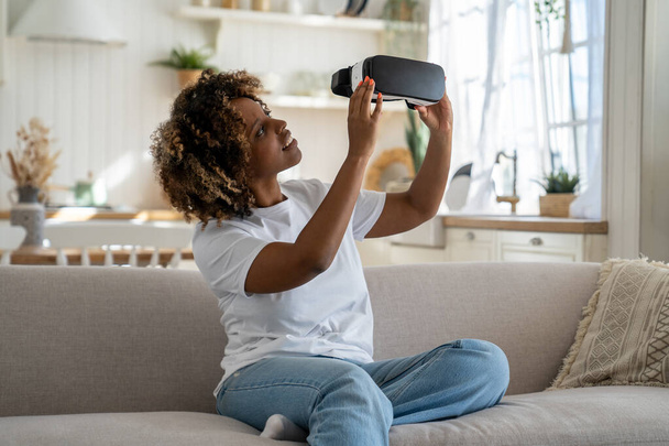Захоплена афро-американська жінка, дивлячись на свій перший VR навушники і відчуваючи себе в захваті, щоб зануритися, сидячи на дивані в вітальні. Цікава чорна леді геймер тримає футуристичні окуляри віртуальної реальності - Фото, зображення