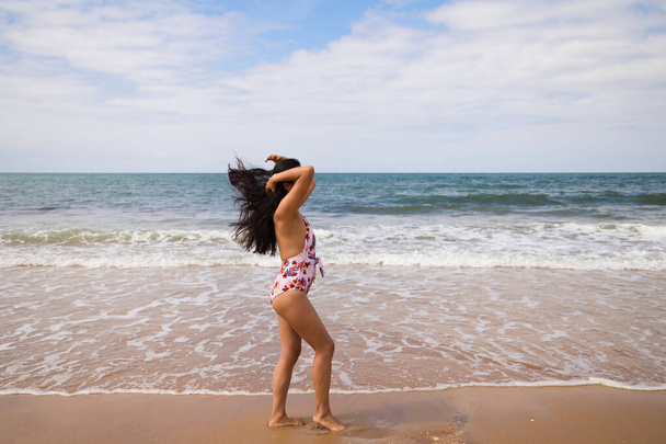 Beautiful and young latin woman is walking along the shore of the beach and posing for photos. Beautiful woman is on holiday on a paradise beach. Holiday and travel concept. - 写真・画像