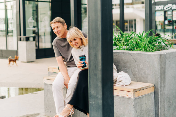 An adult mature happy couple in love sitting on bench outdoors in city street park. A blonde caucasian man and woman spend time together and drinking coffee. Senior wife and husband walking outside - Φωτογραφία, εικόνα