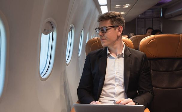 Zakenman in formele kleding en bril die tijdens de vlucht uit het raam van het vliegtuig kijkt terwijl hij met een laptop werkt. Internetverbinding aan boord service en zakenreis concept - Foto, afbeelding