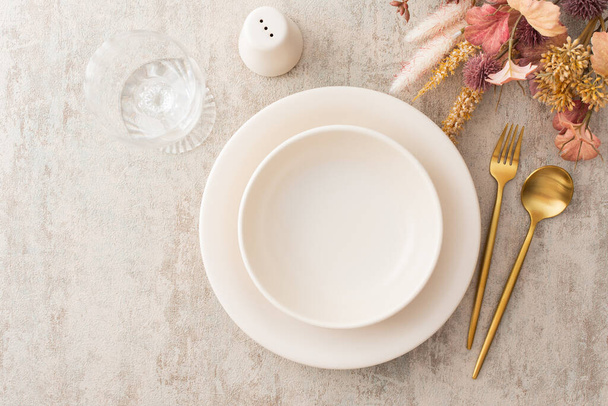 Cadre de table, assiette et couverts dorés sur fond marron, verre à vin vide, vue de dessus de la table servie décorée de fleurs sèches - Photo, image