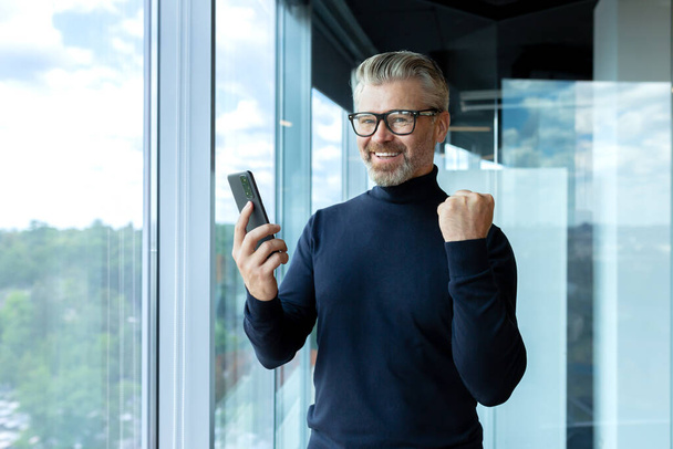Portrait of successful boss, businessman celebrating victory success and good achievement results, man smiling and looking at camera, got win online in phone, employee inside office at workplace. - Foto, Imagen