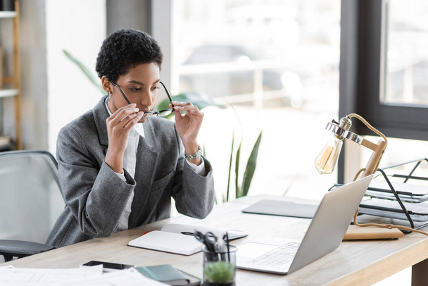 stylish african american businesswoman putting on eyeglasses while sitting near laptop and blank notebook in office - Photo, Image