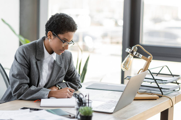 Gözlüklü genç Afrikalı Amerikalı iş kadını ofiste çalışırken dizüstü bilgisayara bakıyor ve not defterine yazıyor. - Fotoğraf, Görsel