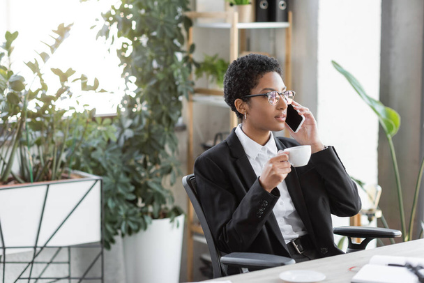 junge afrikanisch-amerikanische Geschäftsfrau mit Kaffeetasse telefoniert in modernem Büro - Foto, Bild