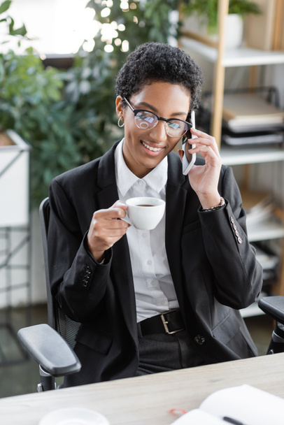 happy african american businesswoman in eyeglasses and black blazer sitting with coffee cup and talking on cellphone in office - Fotó, kép