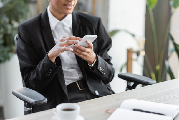 gedeeltelijke weergave van Afrikaanse Amerikaanse zakenvrouw in zwarte blazer messaging op mobiele telefoon in de buurt wazig koffiekopje in het kantoor - Foto, afbeelding