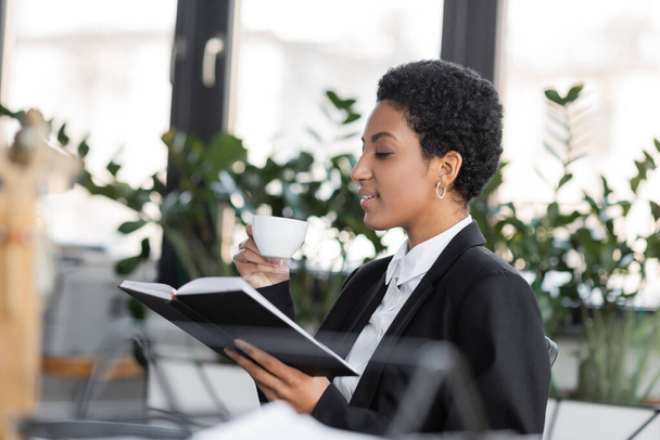side view of african american businesswoman sitting with notebook and coffee cup in office - Фото, изображение