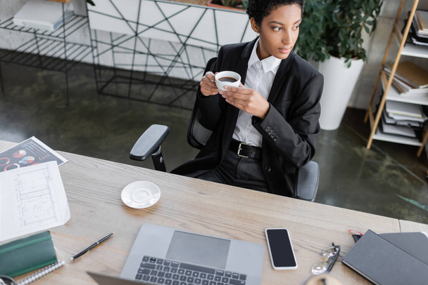 Siyahi blazer ceketli dalgın Afro-Amerikan iş kadını kahve fincanı ile oturuyor ve ofisteki çalışma masasının yakınına bakıyor. - Fotoğraf, Görsel