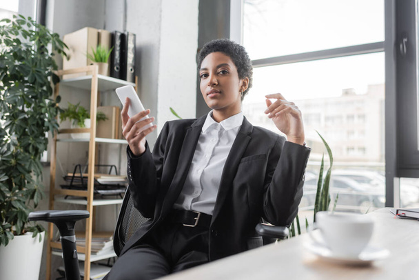 young african american businesswoman in black suit holding mobile phone and looking at camera near blurred coffee cup in office - Фото, зображення