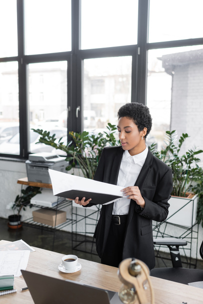 stylish african american businesswoman holding folder with documents while standing near coffee cup on work desk in office - Foto, imagen