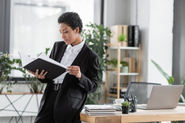 jong Afrikaans Amerikaans zakenvrouw op zoek naar documenten in map terwijl staan in de buurt van bureau met laptop en notebooks in office - Foto, afbeelding