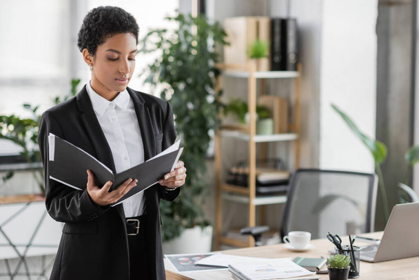 african american businesswoman in black suit looking at folder with papers while standing in office - Foto, Imagem