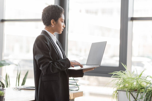 side view of african american businesswoman in black suit using laptop with blank screen while standing in office - Photo, image
