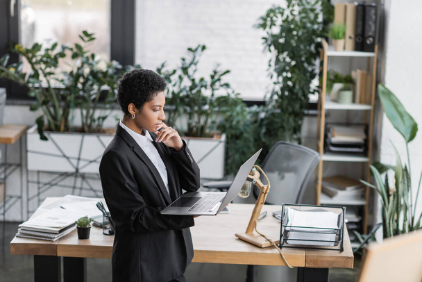 side view of thoughtful african american businesswoman standing in modern office and looking at laptop - 写真・画像