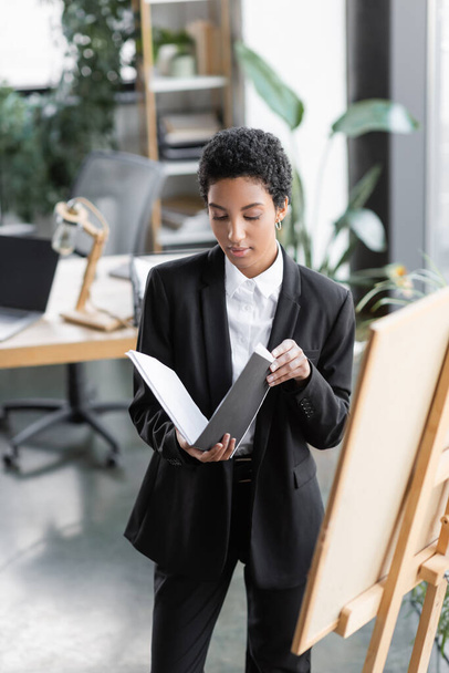 africký americký podnikateležena v černém obleku při pohledu na dokumenty ve složce, zatímco stojí v blízkosti poznámky palubě v úřadu - Fotografie, Obrázek