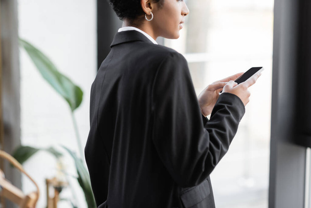 vista parcial de la joven mujer de negocios afroamericana en blazer negro charlando en el teléfono móvil en la oficina - Foto, Imagen