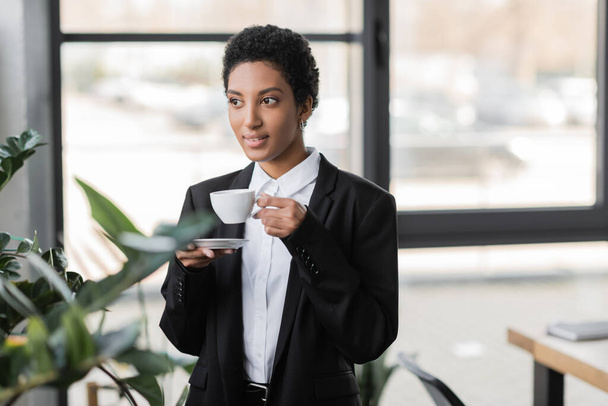 smiling african american businesswoman in black blazer standing with cup of coffee and looking away in office - Foto, Imagem