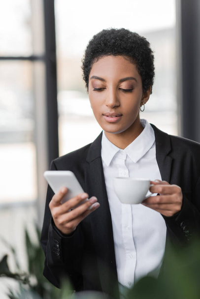 mladá a stylová africká americká podnikatelka v černém blejzru pomocí mobilního telefonu při stání s šálkem kávy v kanceláři - Fotografie, Obrázek
