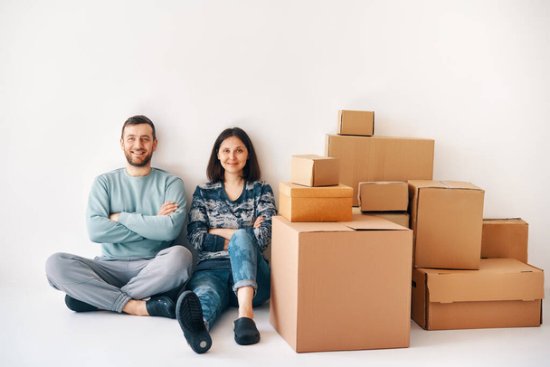 Smiling happy couple sitting on floor after the moving to new house with cardboard boxes on background. Real estate, new apartment concept - Foto, imagen