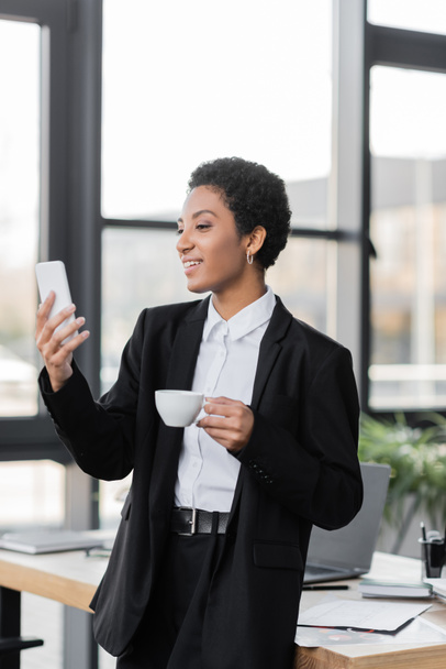 vidám afro-amerikai üzletasszony kezében egy csésze kávét, és nézte okostelefon az irodában - Fotó, kép