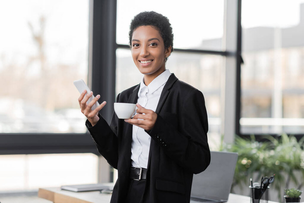 pleased african american businesswoman with mobile phone and coffee cup looking at camera in office - Foto, Imagem