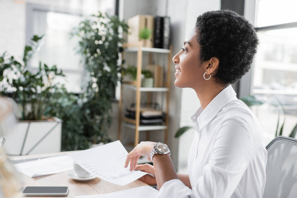 side view of african american businesswoman smiling and looking away while working with papers in office - Foto, afbeelding