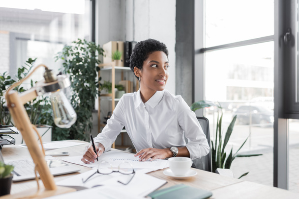 happy african american businesswoman sitting with pen near documents and looking away in office - 写真・画像