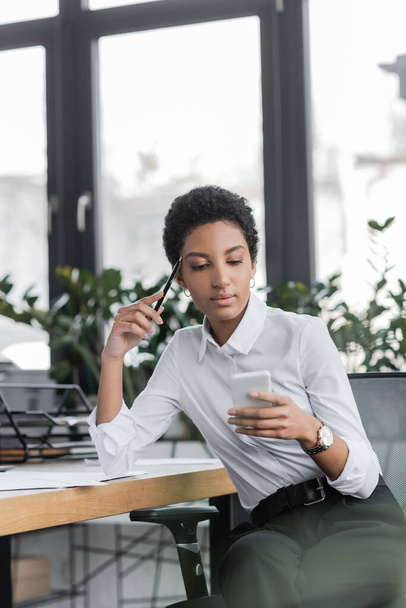 nachdenkliche afrikanisch-amerikanische Geschäftsfrau in weißer Bluse hält Stift in der Hand und blickt im Büro auf ihr Smartphone - Foto, Bild