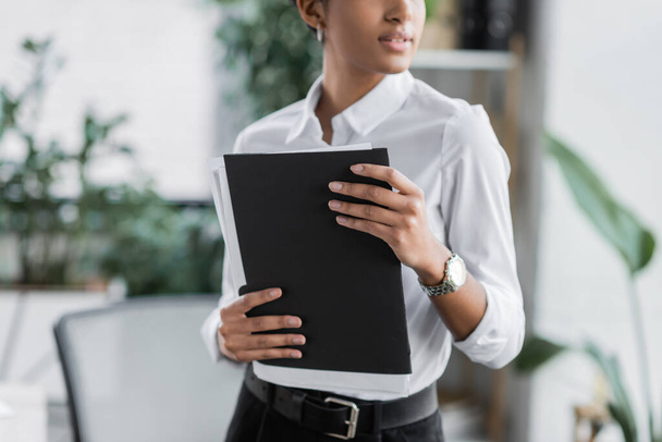 bijgesneden weergave van Afrikaanse Amerikaanse zakenvrouw in witte blouse holding map met documenten in het kantoor - Foto, afbeelding