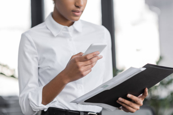 vista recortada de la mujer de negocios afroamericana en blusa blanca que sostiene la carpeta y mensajería en el teléfono inteligente en la oficina - Foto, Imagen