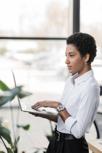 side view of dreamy african american businesswoman in white shirt holding laptop and looking away in office - Foto, immagini