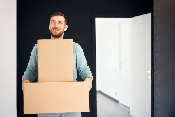 Young smiling man moving into new apartment carrying cardboard boxes. Relocation concept - Valokuva, kuva