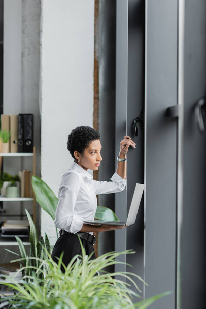 вдумчивая американская бизнесвумен с ноутбуком, смотрящая в сторону окон в современном офисе - Фото, изображение